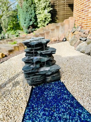 Une cascade artificielle en roche, avec une base en verre bleu, entourée de gravier et d’un mur de briques.