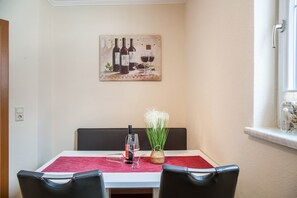 Zona pranzo con tavolo apparecchiato per due, una bottiglia di vino e un vaso con piante verdi.