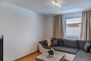 Ein modernes Wohnzimmer mit einem Ecksofa, einem kleinen Tisch mit Pflanze und einem Fenster mit Vorhängen.