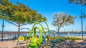 Un parque infantil con un tobogán verde y estructuras para escalar, cerca de un paseo marítimo con un puente al fondo.