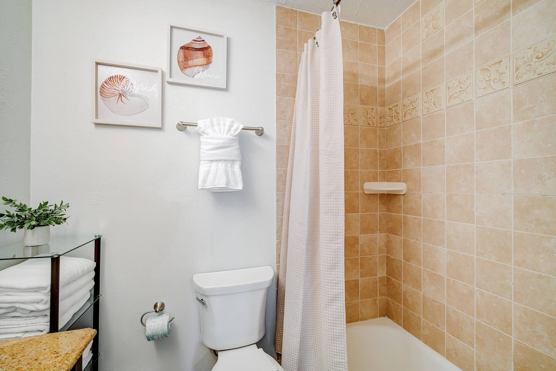 Un baño con inodoro, lavamanos, toallero y ducha con azulejos color crema.