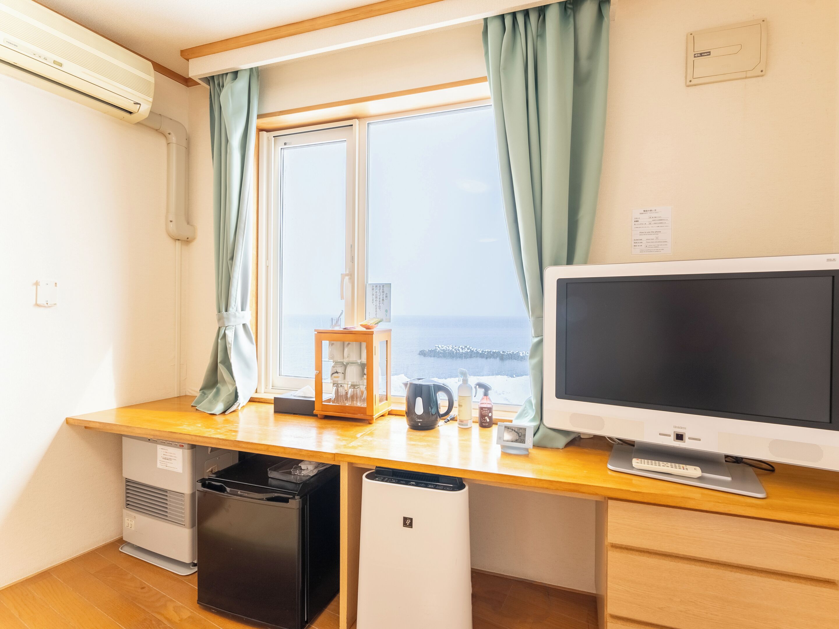 A compact room with a wooden desk, a television, a small refrigerator, and a window with curtains.
