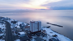 An aerial view of a coastal town with a prominent hotel featuring multiple floors and a flat roof, situated near a calm body of water with a dock extending into it. - Livemax Resort Shiretoko Seafront (Rausu)