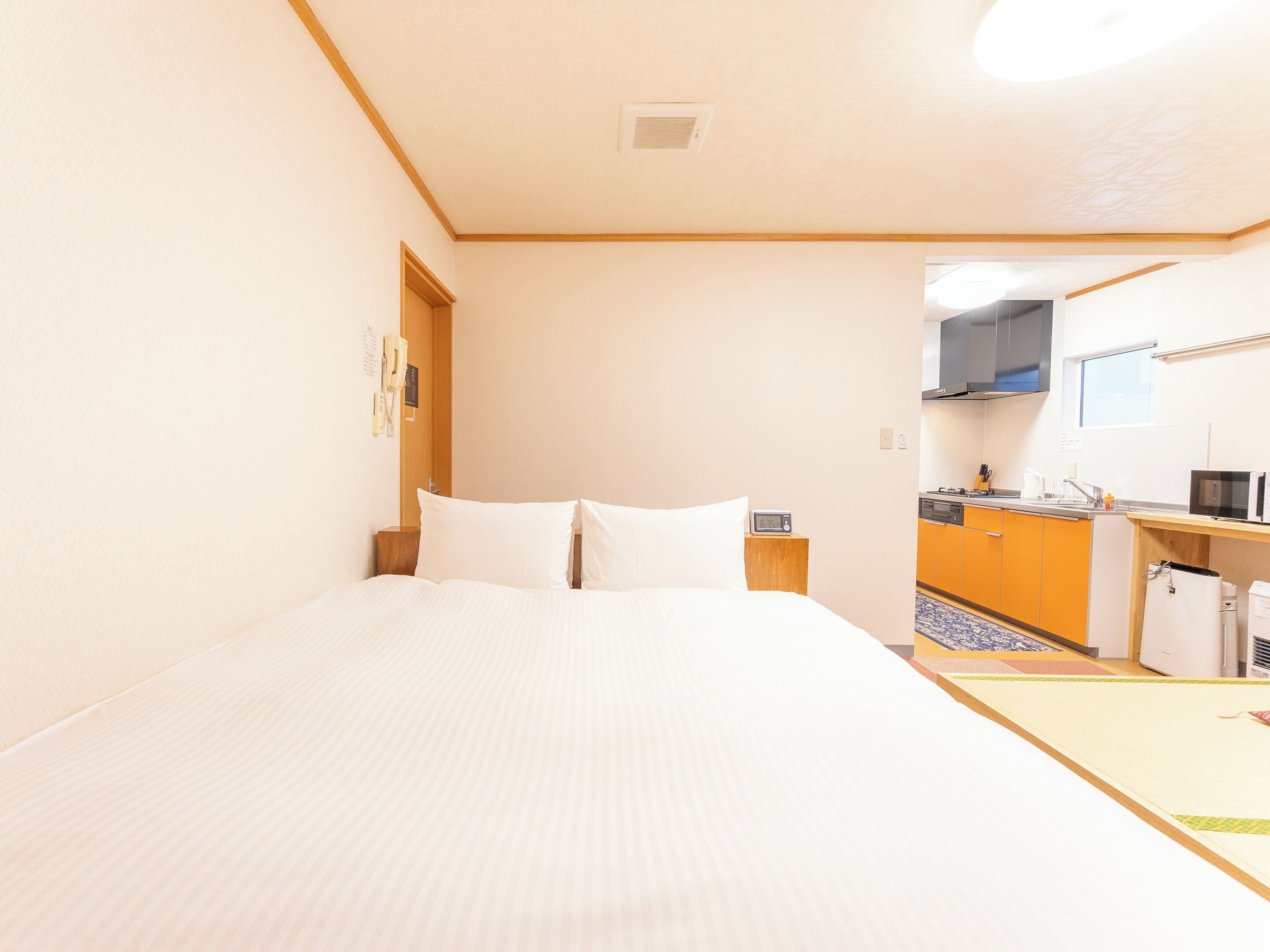 Een hotelkamer met een bed, een houten nachtkastje, een telefoon aan de muur en een kitchenette met een magnetron en spoelbak.