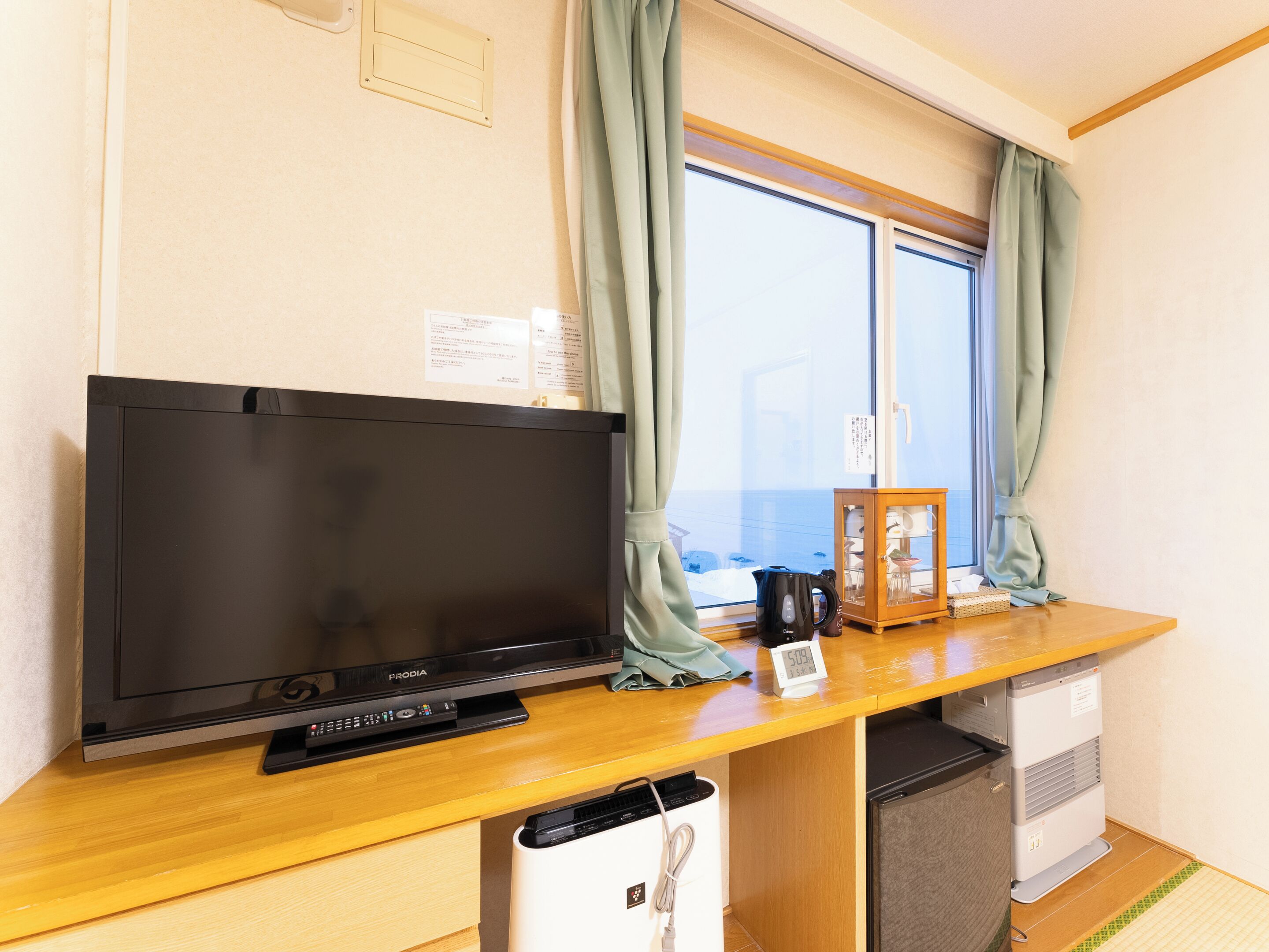 A room with a television, a small refrigerator, a kettle, and a window with curtains.