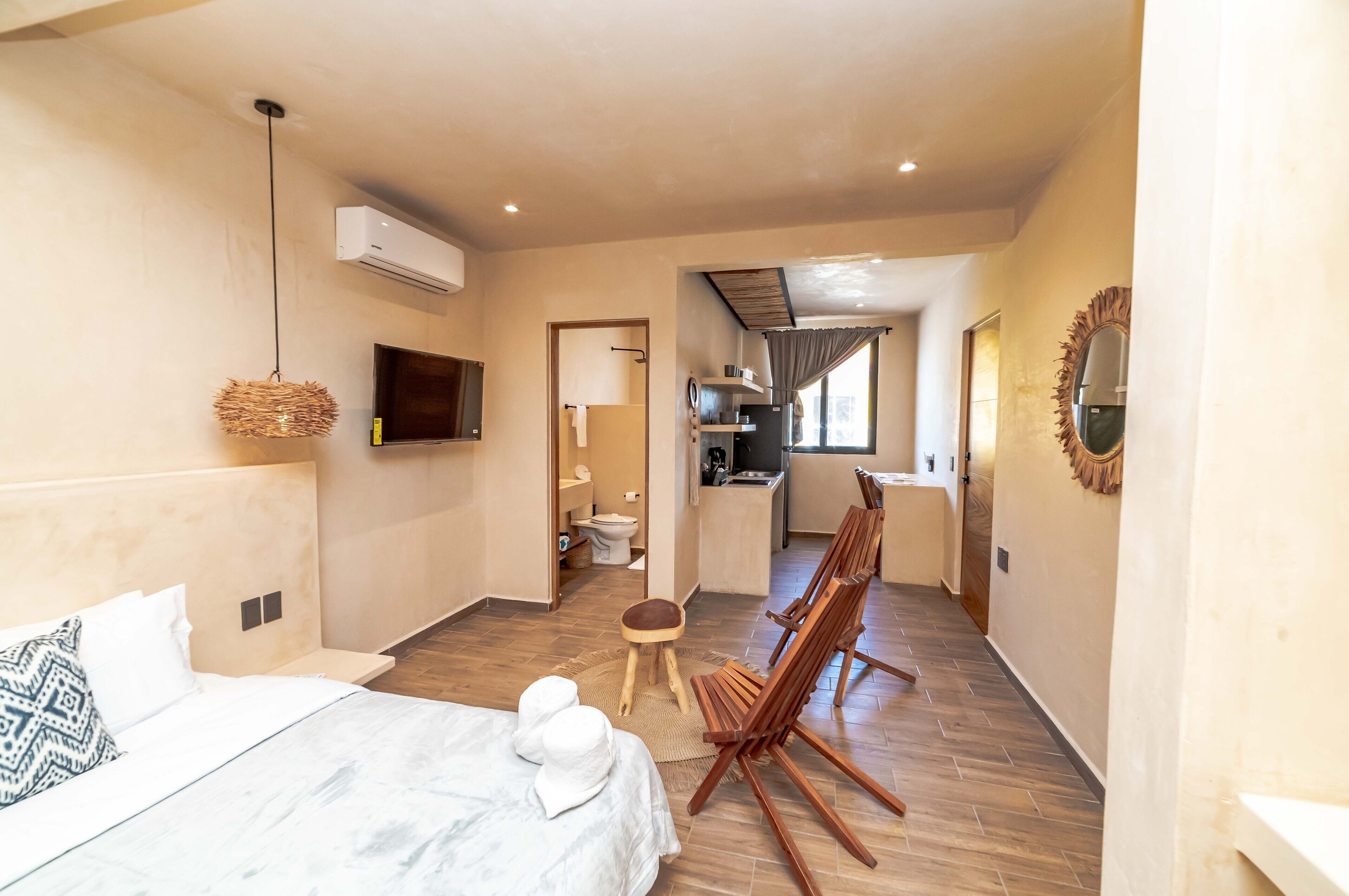 Habitació d'hotel amb un llit, una televisió, una cadira de fusta, un petit tamboret i una zona de cuina al fons.