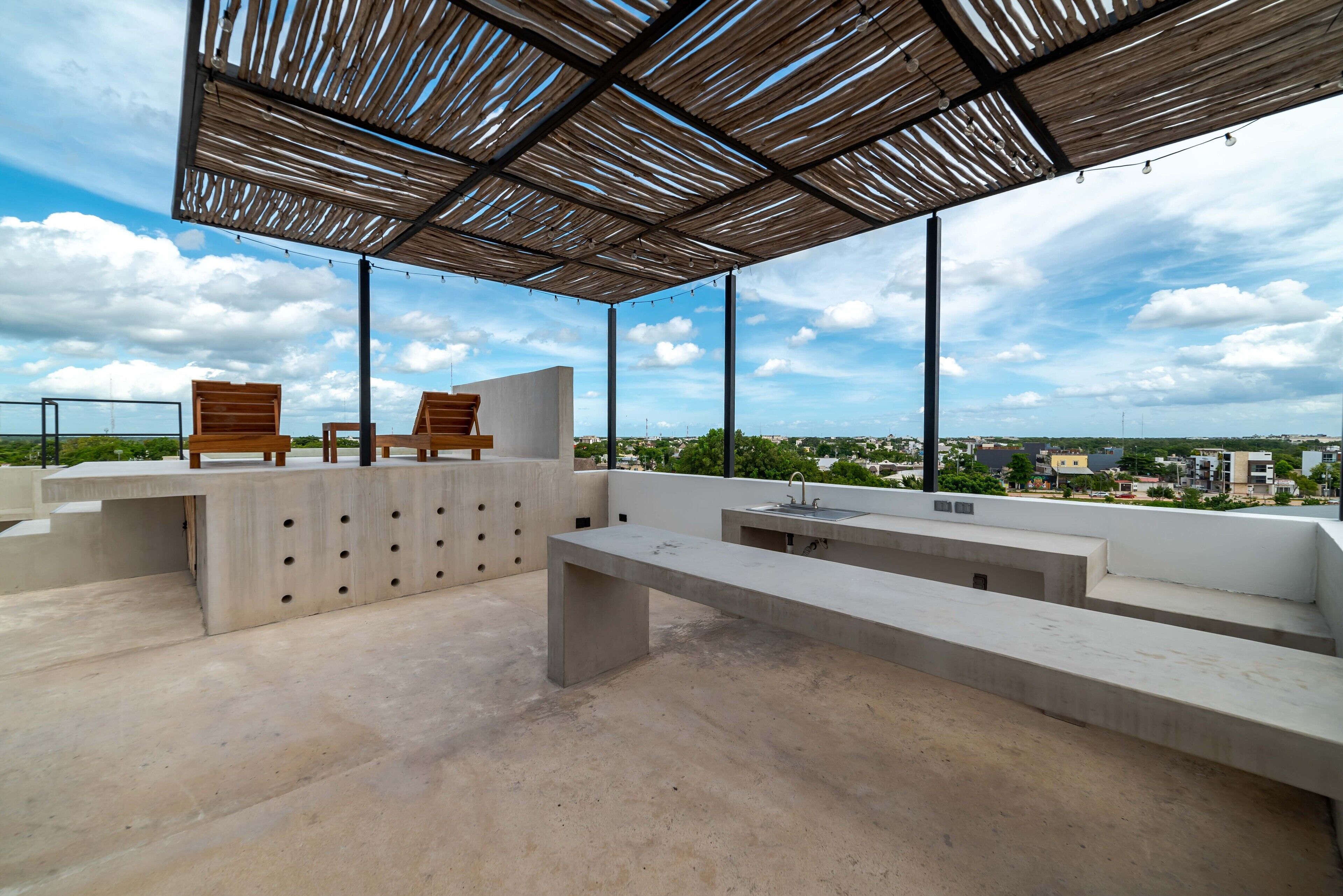 An outdoor terrace with concrete benches and wooden chairs, a metal pergola, and a view of the cityscape.