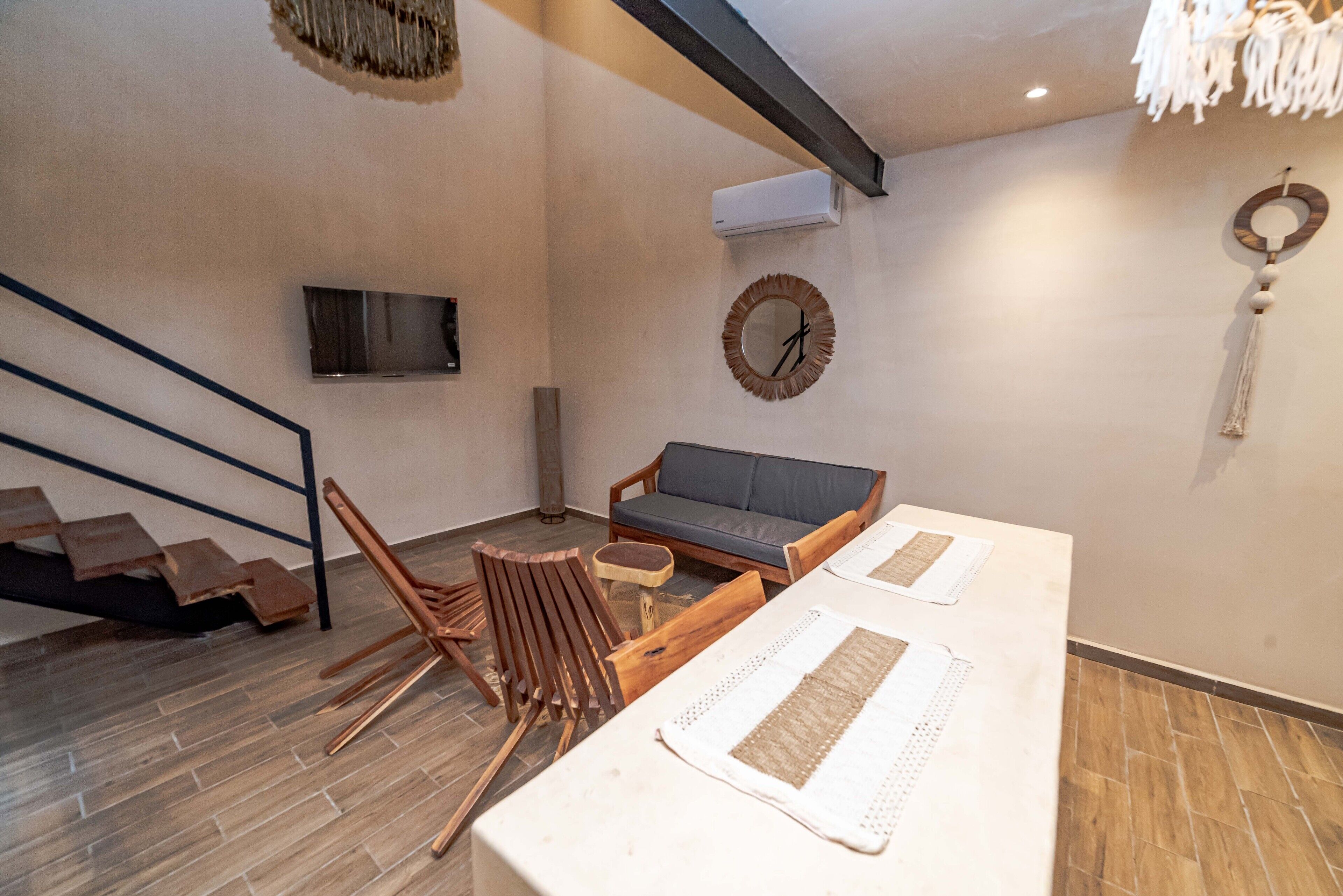 A modern living room with a sofa, coffee table, and chairs. There is a staircase with wooden steps and a metal railing. An air conditioning unit is mounted on the wall.