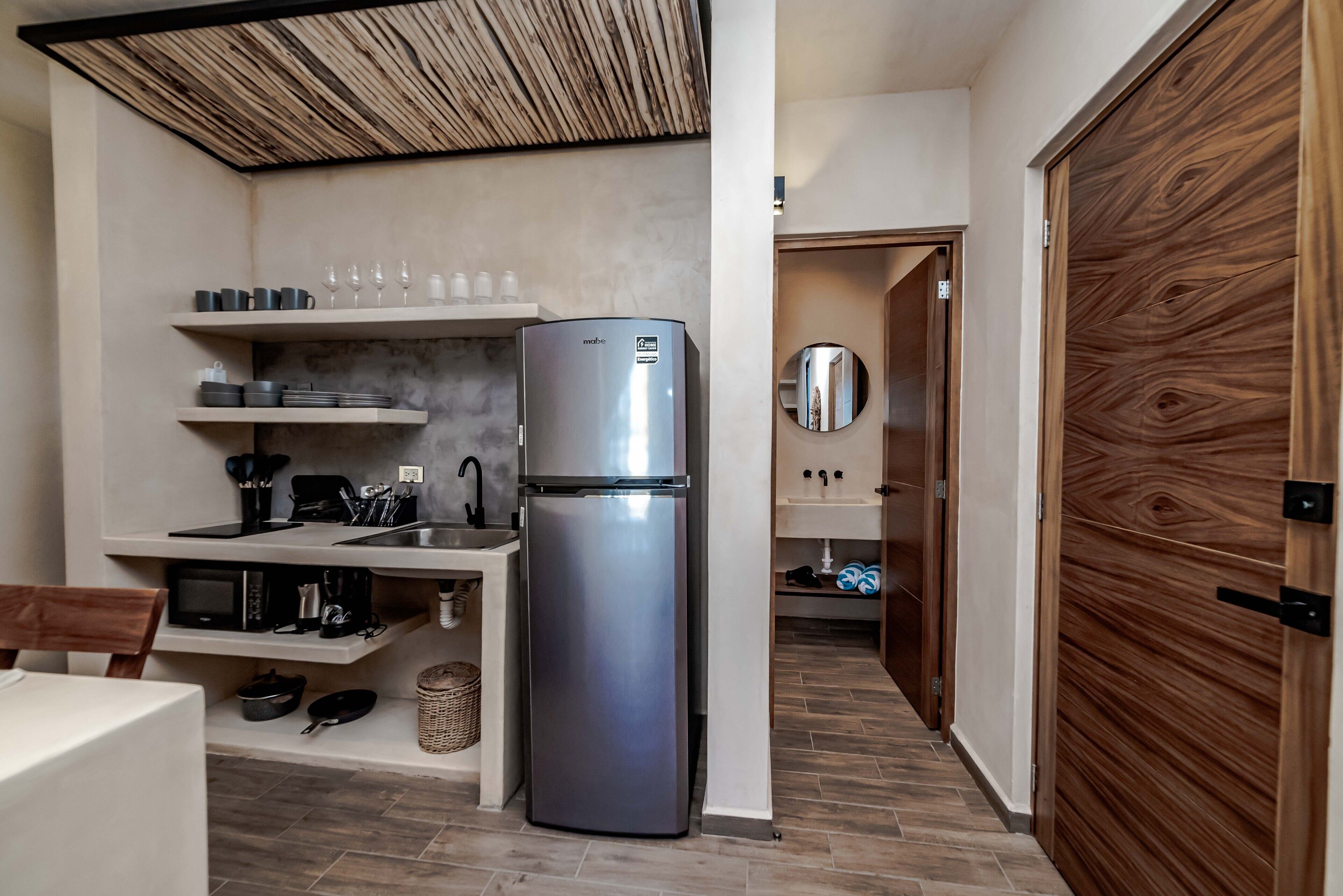 A compact kitchen with a built-in refrigerator, a microwave, and a sink. There are shelves above the counter holding cups and glasses.