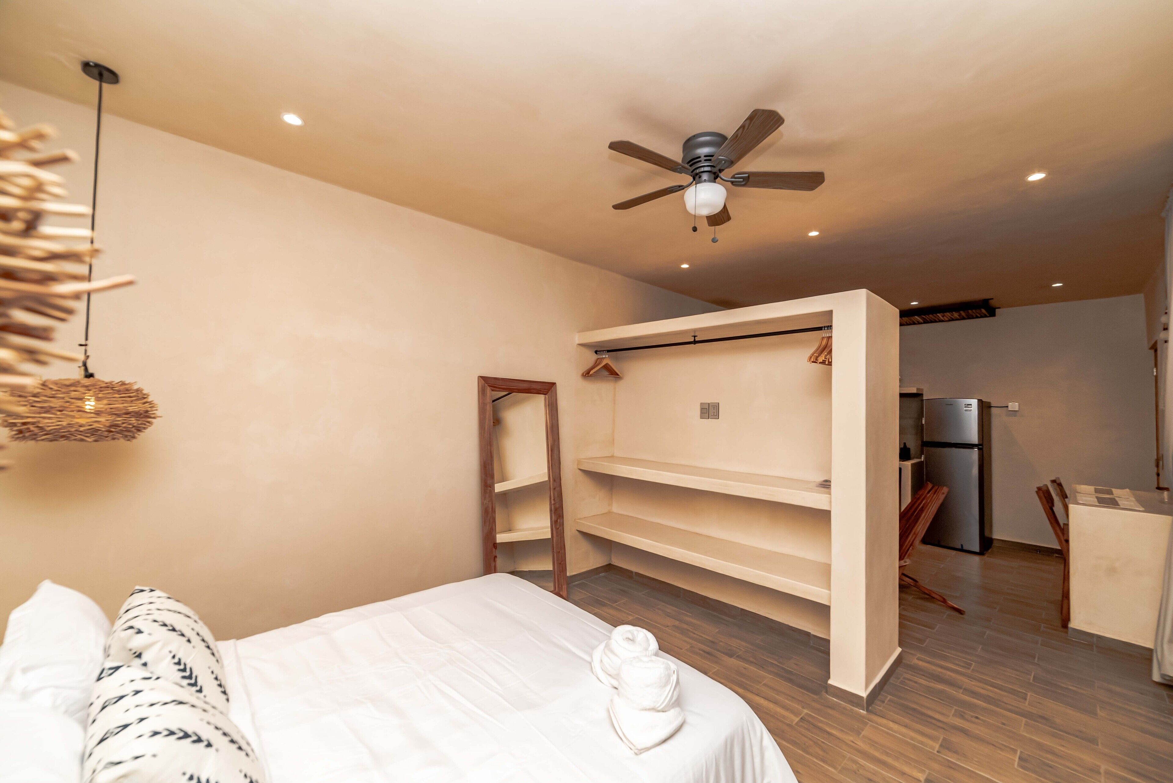 A bedroom with a bed, a ceiling fan, a hanging light, a wooden wardrobe, and a kitchenette with a refrigerator.