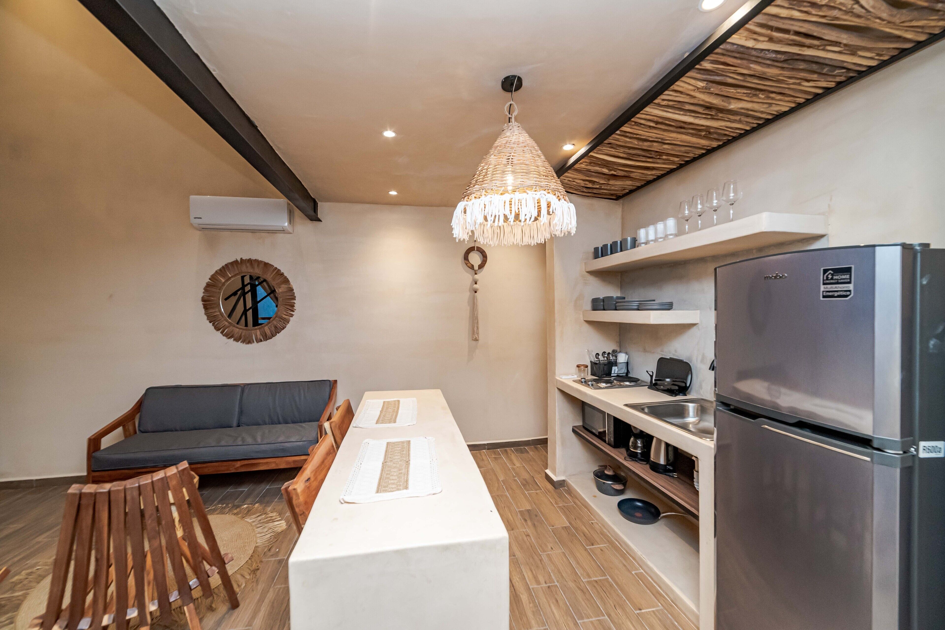 Une cuisine compacte comprenant un réfrigérateur, une table à manger avec deux chaises, un luminaire suspendu et une étagère intégrée pour ranger les ustensiles de cuisine.