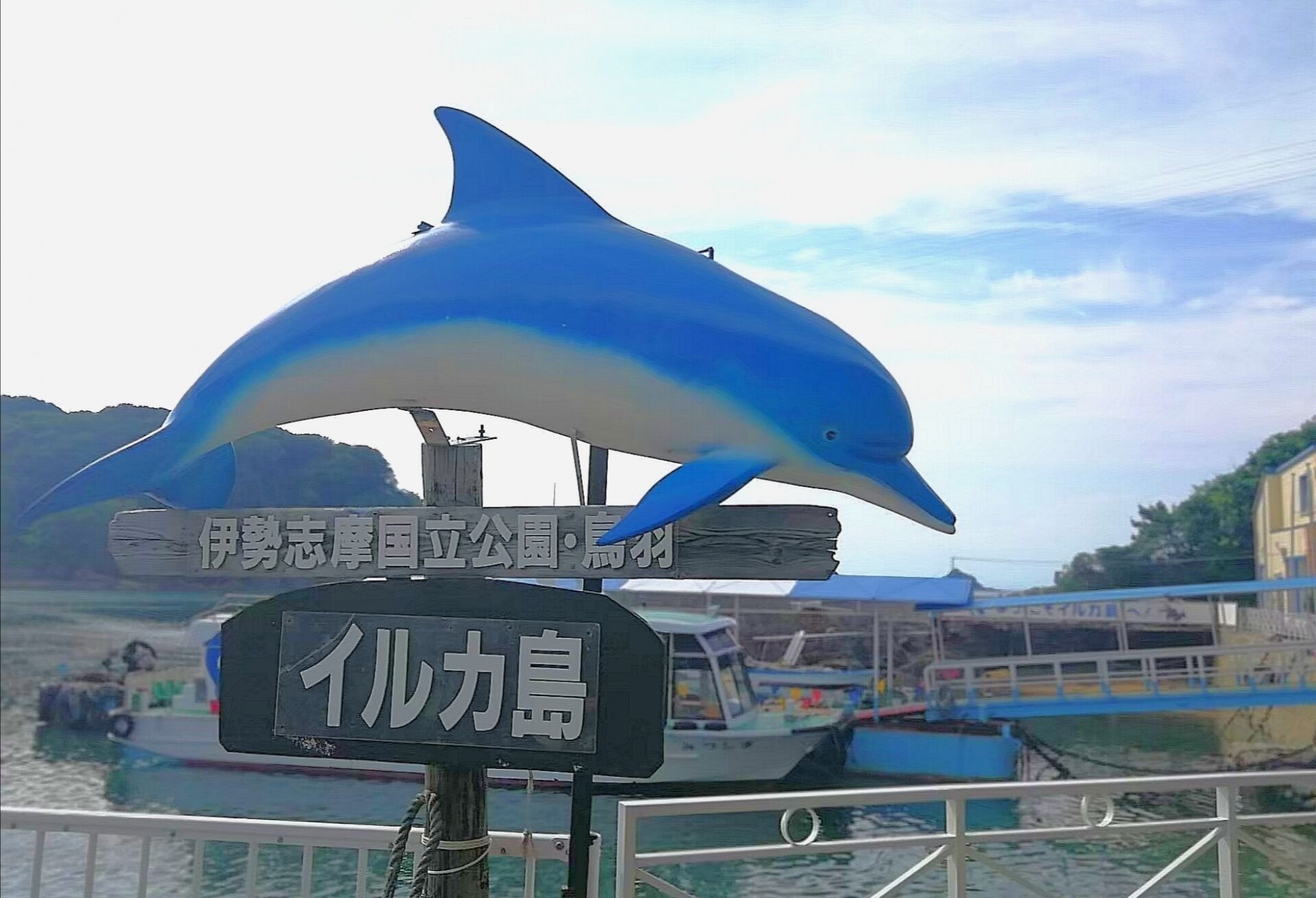 A dolphin sculpture above a sign with Japanese text, a boat in the background, and a railing in the foreground.
