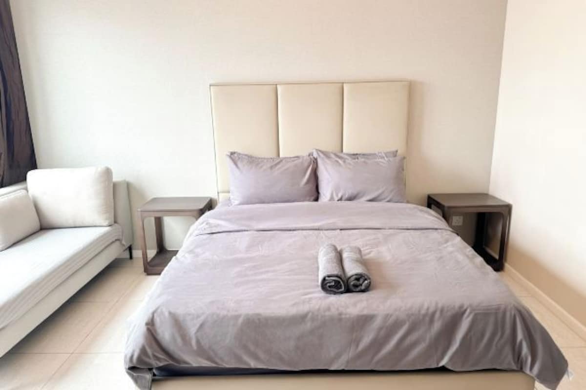 A neatly made bed with a headboard, two pillows, and rolled towels on the bed.