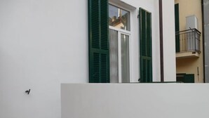 Une pièce avec un mur blanc, une fenêtre avec des volets verts et un balcon visible, doté d’une balustrade.