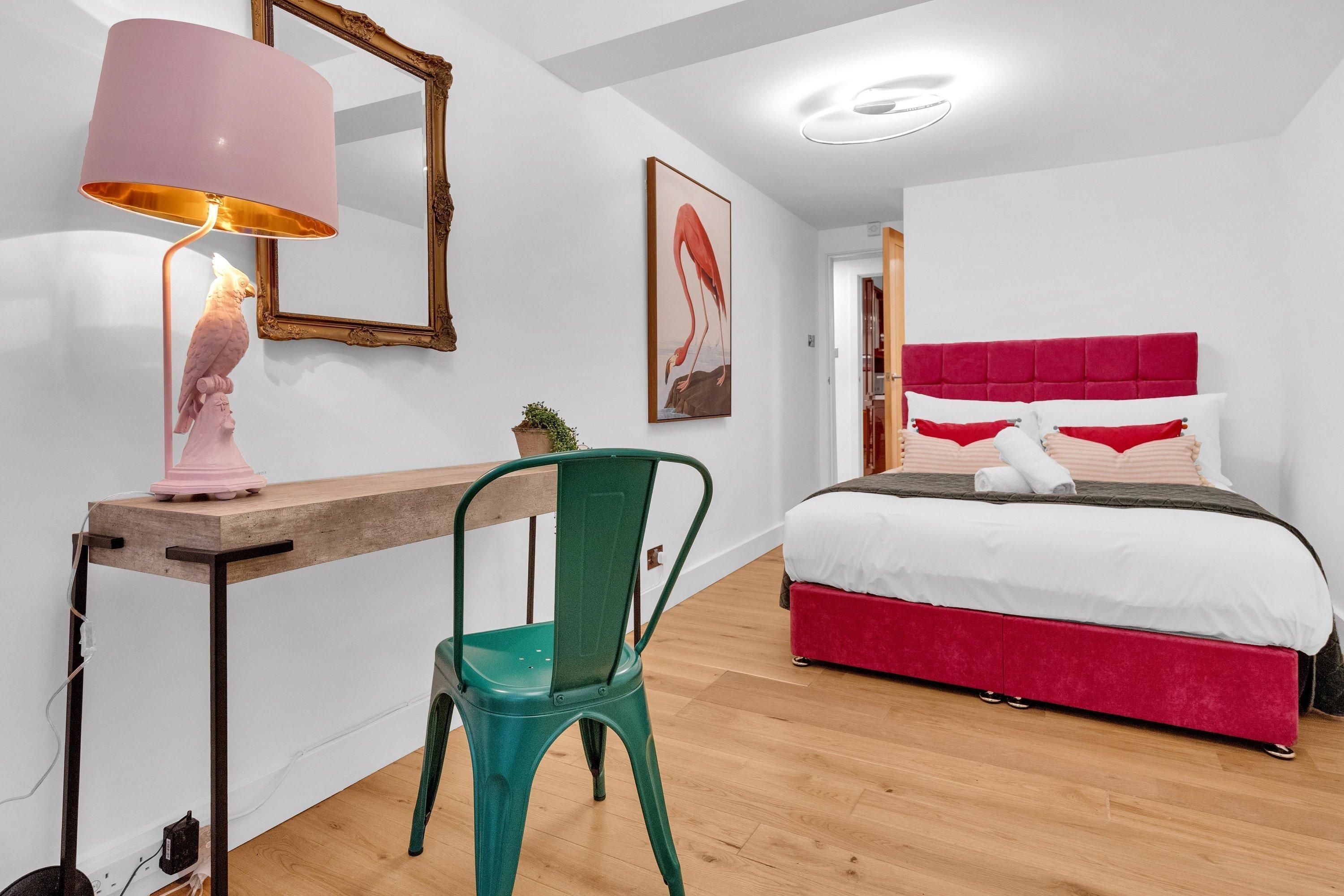 A bedroom with a bed, a desk with a lamp, a chair, a mirror, and a painting on the wall.
