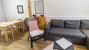 Un salon moderne avec un canapé gris, un fauteuil noir agrémenté d’un coussin rose, une table à manger en bois avec des chaises, et un tableau encadré accroché au mur.