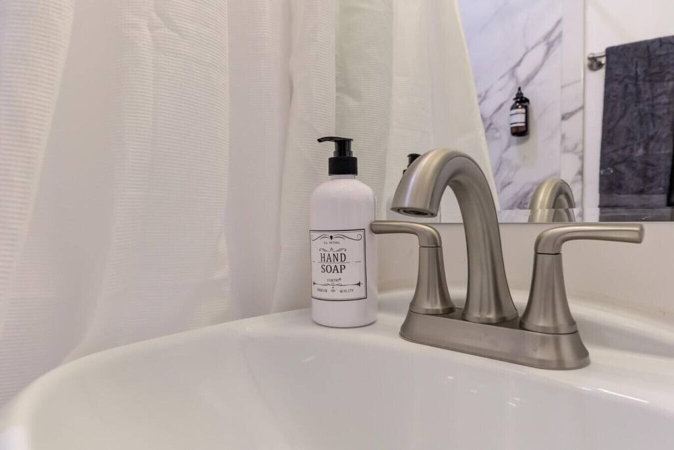 A bathroom with a white sink, a hand soap dispenser, a modern faucet, and a mirror reflecting a marble wall.