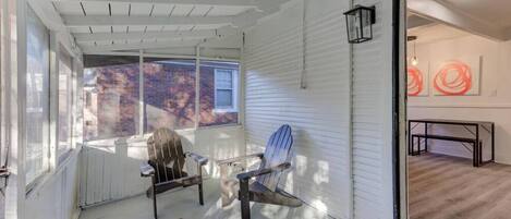A covered porch with two Adirondack chairs, a sliding glass door leading to an interior space, and a view of a brick building.