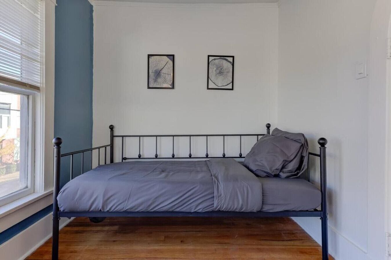 Een eenpersoonsbed met een metalen frame, grijze beddengoed en twee ingelijste kunstwerken aan de muur.