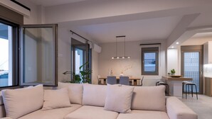 A modern living room with a beige sectional sofa, a wooden coffee table with a pitcher and glass, and a dining area with a table and chairs. - Leon Luxury Home in Rethymno (Rethymno)