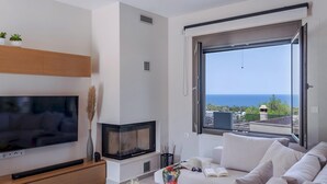 A modern living room with a flat-screen TV, a fireplace, a sofa, and a large window offering a view of the sea. - Leon Luxury Home in Rethymno (Rethymno)