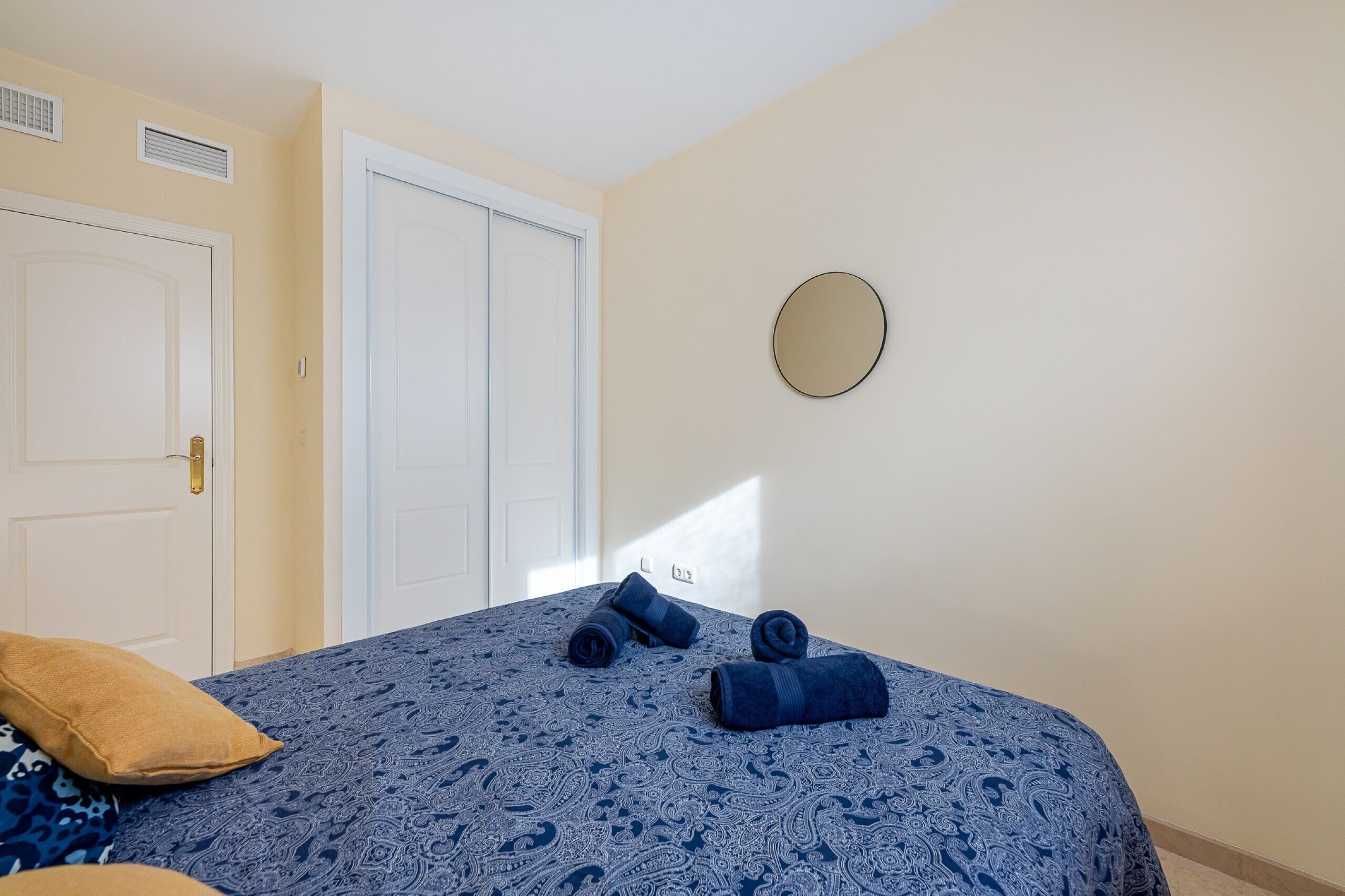 A bedroom with a bed, two rolled towels, a round mirror, and a door.