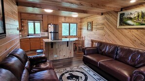 Un intérieur de chalet chaleureux, avec un revêtement mural en bois, une kitchenette, un coin repas et un salon doté de canapés en cuir.