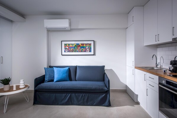 A modern living room with a dark blue sofa, a small side table with decorative items, a framed colorful artwork on the wall, and a kitchen area with white cabinets and a wooden countertop. - Athinais Apartments (Heraklion)