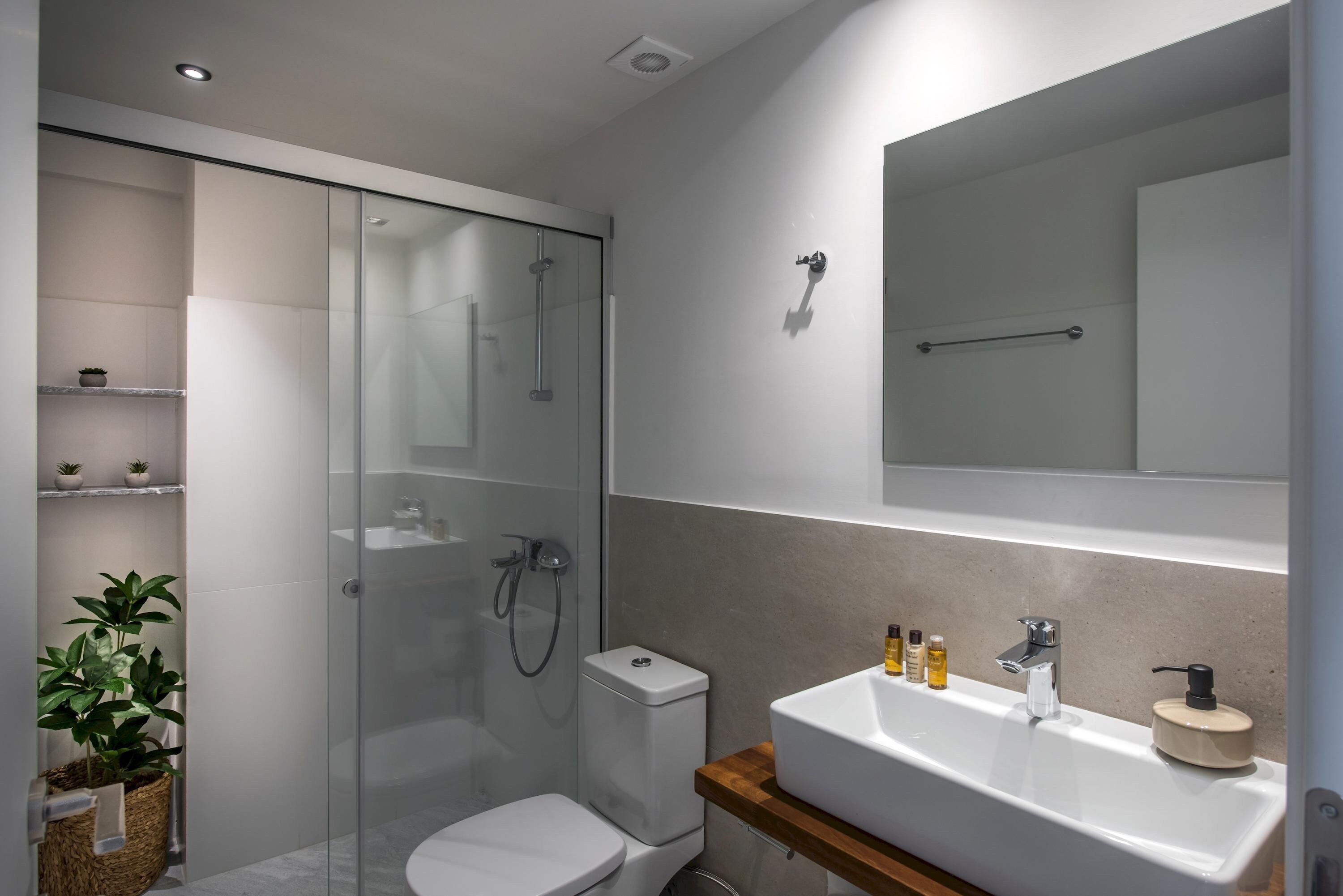 Un baño moderno con ducha de vidrio, inodoro, lavamanos con artículos de aseo y un espejo sobre el lavamanos.