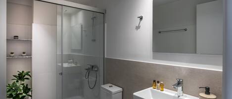 Un baño moderno con ducha de vidrio, inodoro, lavamanos con artículos de aseo y un espejo sobre el lavamanos.