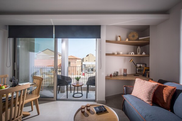 A modern living room with a glass door leading to a balcony, a wooden dining table, and a blue sofa with patterned cushions. - Athinais Apartments (Heraklion)