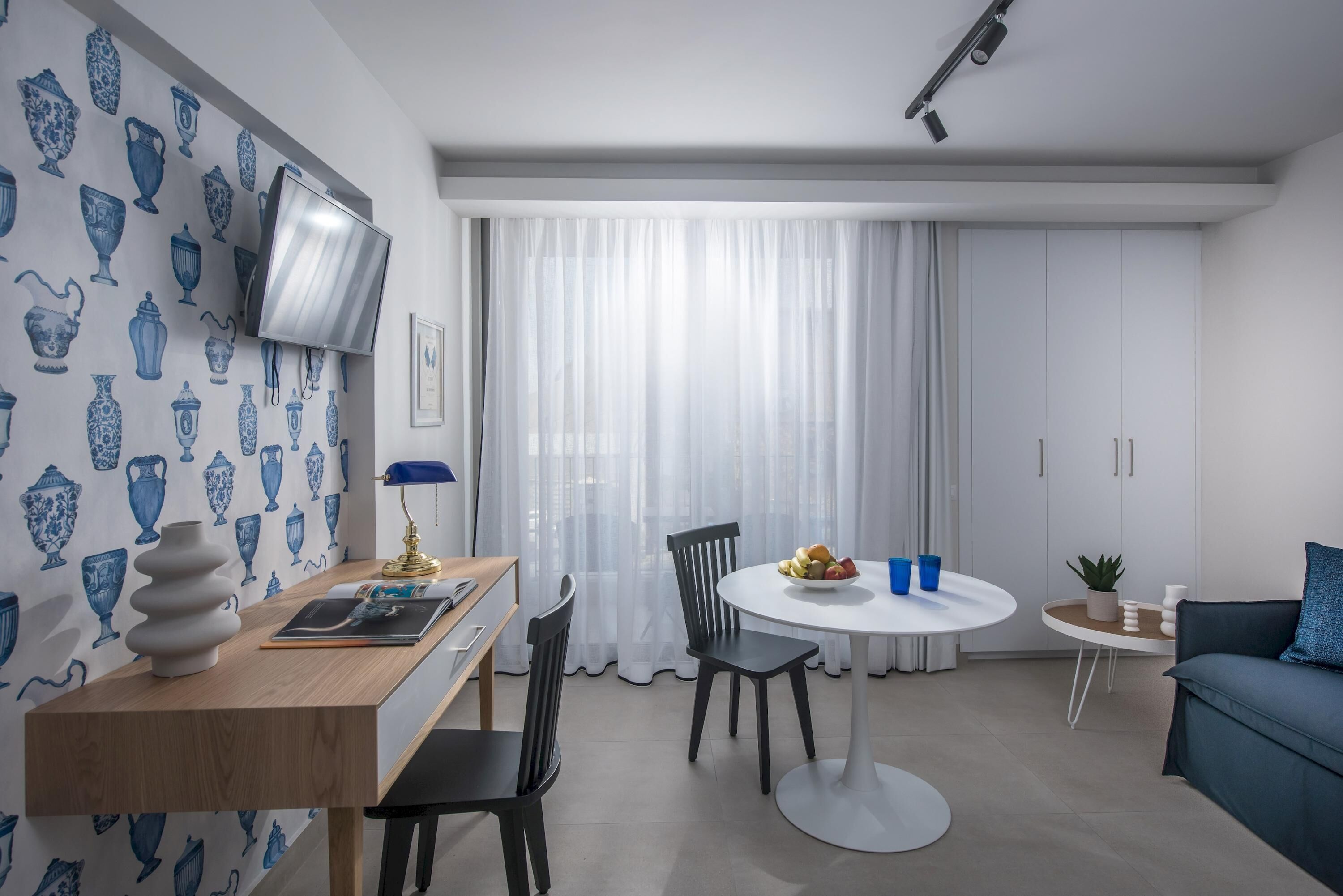 A modern living room with a dining area, featuring a flat-screen TV mounted on the wall, a round dining table with chairs, a blue sofa, and a small side table with a plant.