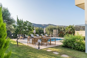 A pool area with lounge chairs and umbrellas, surrounded by greenery and a view of hills.