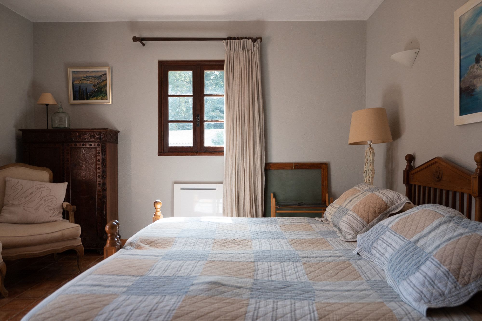A bedroom with a large bed, a wooden chair, a dresser, a window with curtains, and a painting on the wall.