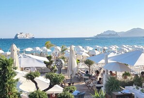 A beachside restaurant with outdoor seating, umbrellas, and a view of the sea and cruise ships.
