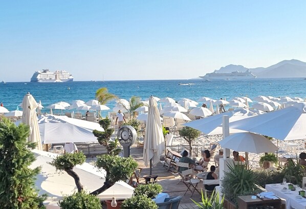 A beachside restaurant with outdoor seating, umbrellas, and a view of the sea and cruise ships.