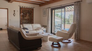Un salon comprenant un canapé, un fauteuil, un pouf et une table basse. On y trouve une tapisserie au mur, une fenêtre avec des rideaux et une vue sur l’extérieur.