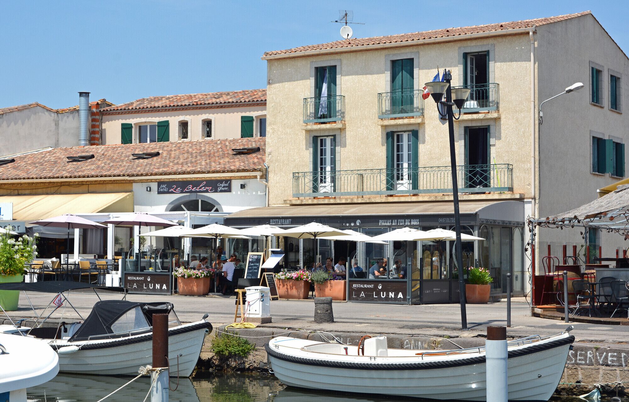 Un restaurante frente al agua con terraza, barcos amarrados cerca y un edificio con balcones y señalización.