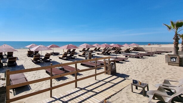 Un hôtel situé en bord de mer, avec des rangées de chaises longues et de parasols, une terrasse en bois et une vue dégagée sur l’océan.