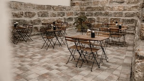 An outdoor seating area with wooden tables and chairs, stone walls, and a tiled floor.