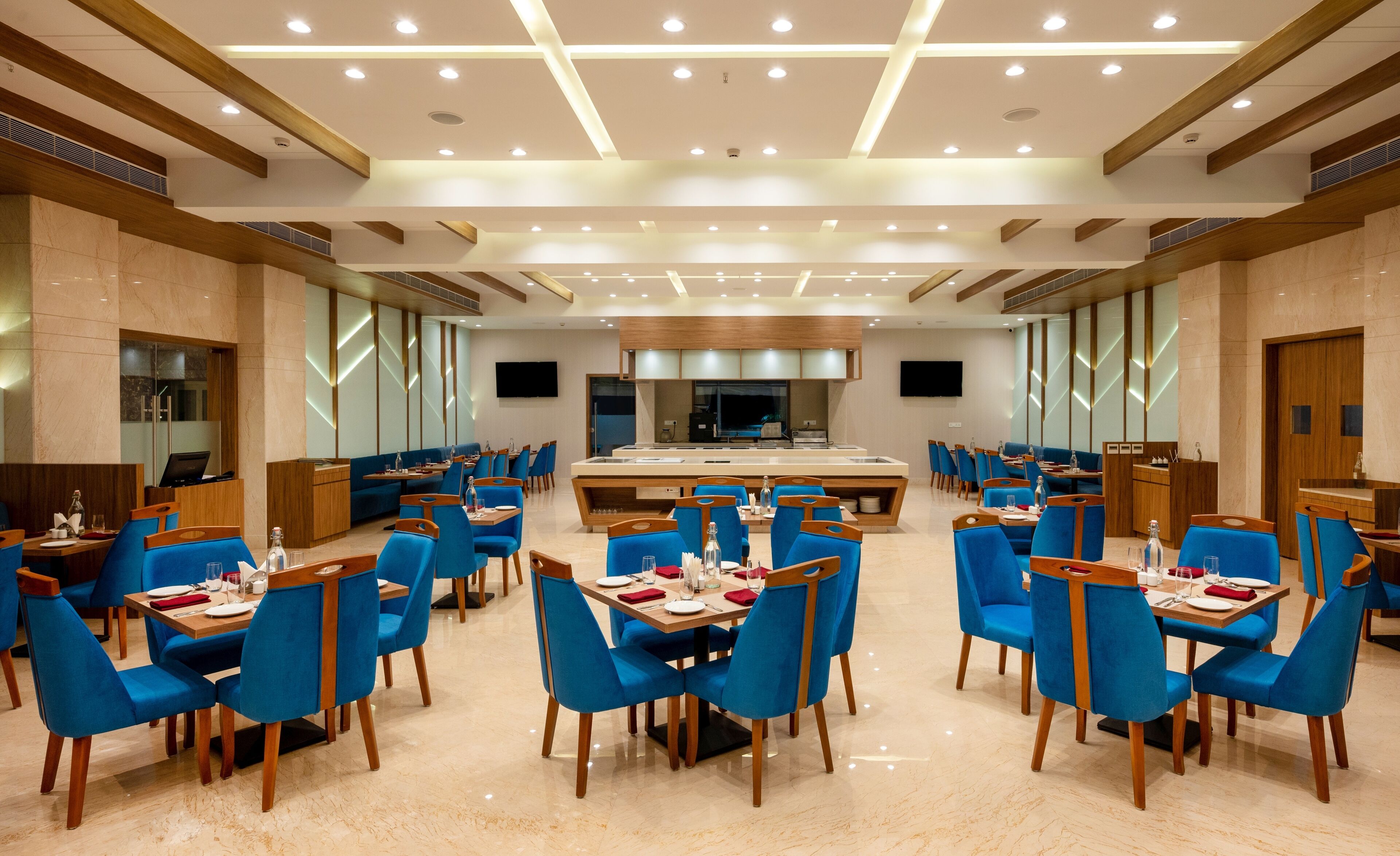 Spaziosa sala da pranzo di un hotel con numerosi tavoli e sedie blu, caratterizzata da un'isola centrale con un lavandino e un televisore a schermo piatto montato a parete.