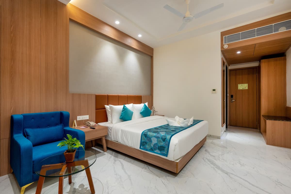Quarto de hotel moderno com uma cama grande, uma poltrona azul, uma pequena mesa com uma planta e um ventilador de teto.