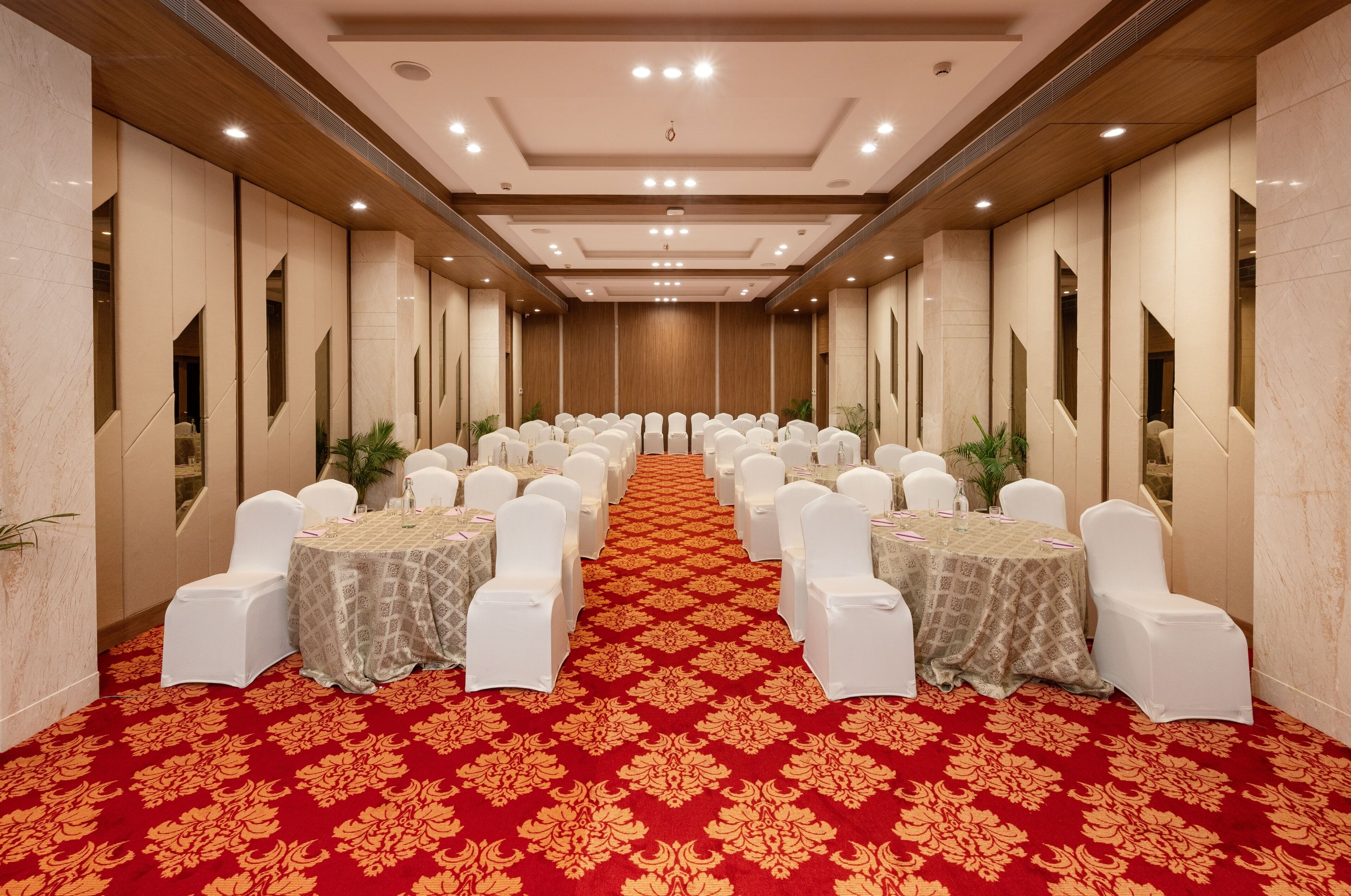 A long, narrow hotel room with rows of white chairs and tables, a red carpet, and wooden paneling on the walls.