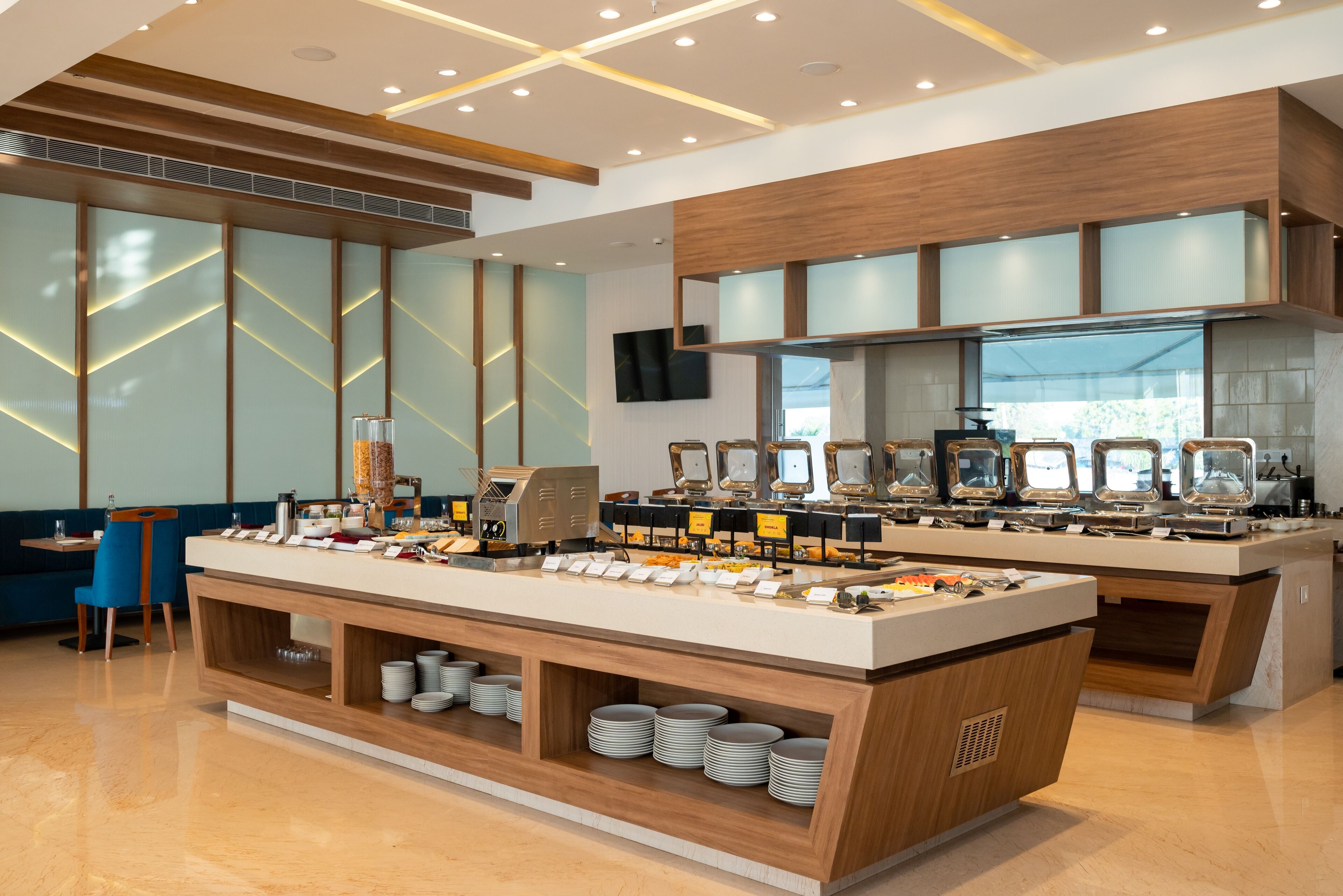 A buffet setup with multiple serving stations, a seating area with blue chairs, and a modern interior design with wooden accents and a tiled backsplash.