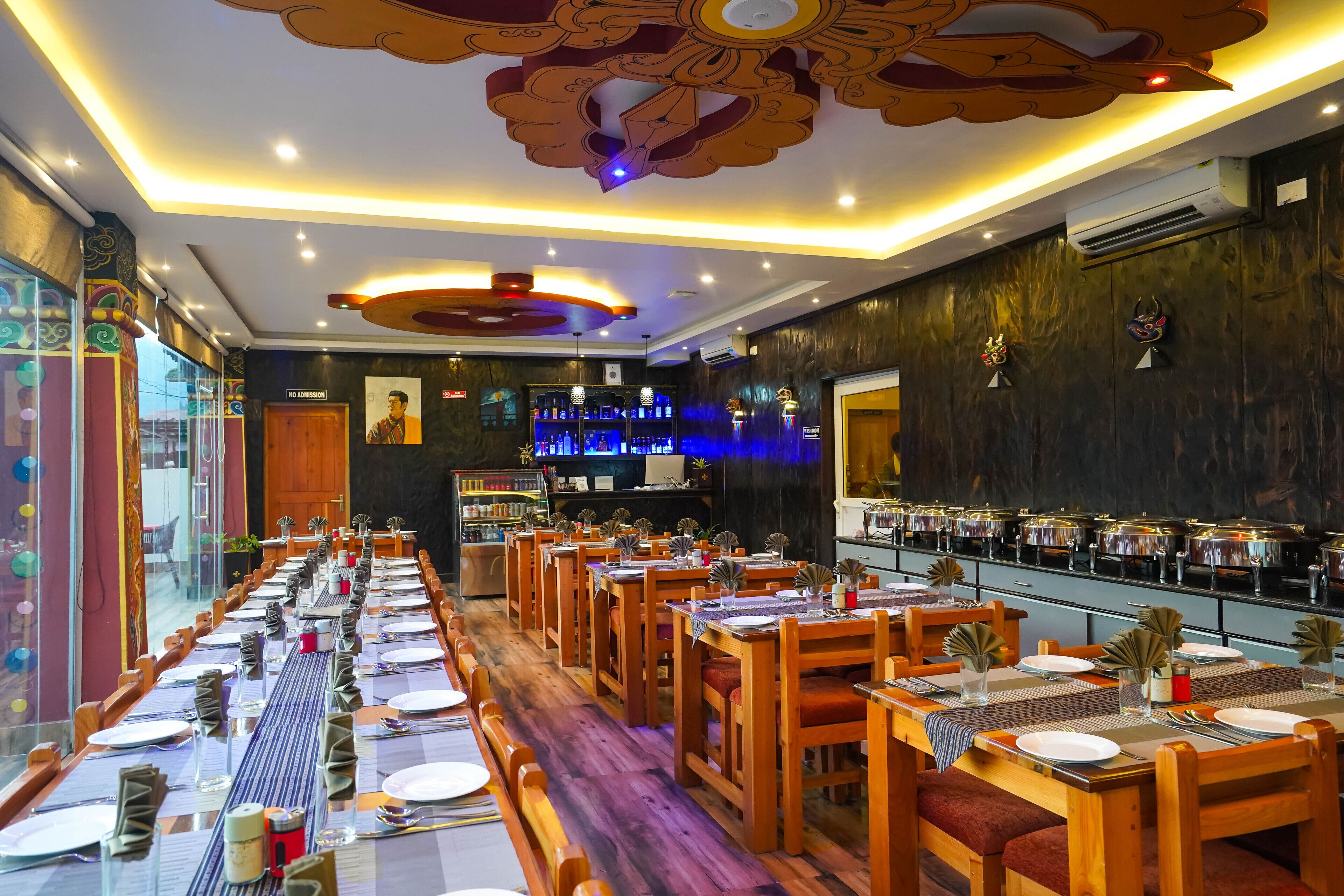 Een restaurant met lange houten tafels, rode stoelen met kussens en een bar met flessen.