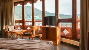 A hotel room with a bed, wooden furniture, a TV on a wooden stand, and large windows offering a view of a mountainous landscape. - Hotel Gakyidiana (Paro)
