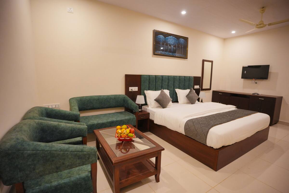 A hotel room with a large bed, a green sofa, a glass-top coffee table with fruit, a TV, and a ceiling fan.