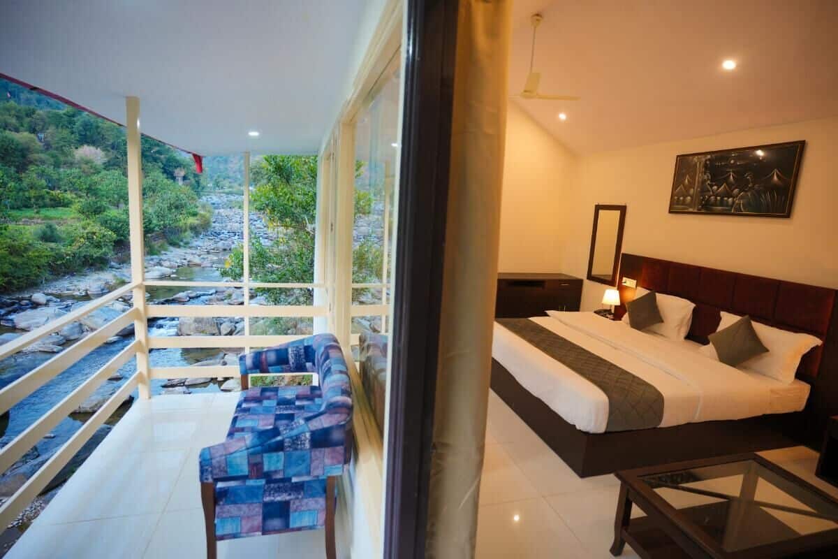 A bedroom with a large bed, a nightstand, a bedside table, a chair, and a view of a river and greenery through large windows.