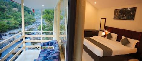A bedroom with a large bed, a nightstand, a bedside table, a chair, and a view of a river and greenery through large windows.