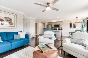 Amplia sala de estar con un sofá azul, un sillón blanco y una mesa de centro. Se observa un ventilador de techo, una lámpara de araña y una cocina al fondo.